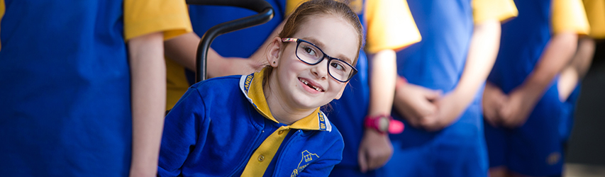 This image is a photograph of a young girl who wears a blue school uniform with yellow trim, glasses, and a beautiful smile. She is seated in a wheelchair, second in a line of student colleagues, and leaning to her left to peer around the person in front of her. Her gaze is drawn by someone on the photographer’s left.